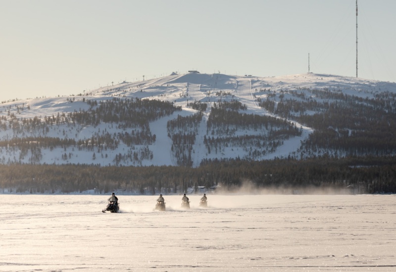 People riding snow scooters, with a large hill coated in snow and covered in trees behind them.