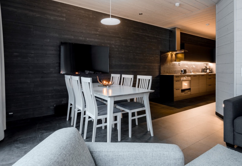 A table with 3 chairs on each sides standing in front of a TV. A cozy kitchen in the background.