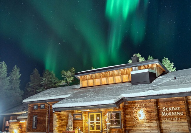 Snow-covered rustic lodge with warm-lit entrance beneath vivid green aurora borealis and starry sky, perfect for northern-lights stays