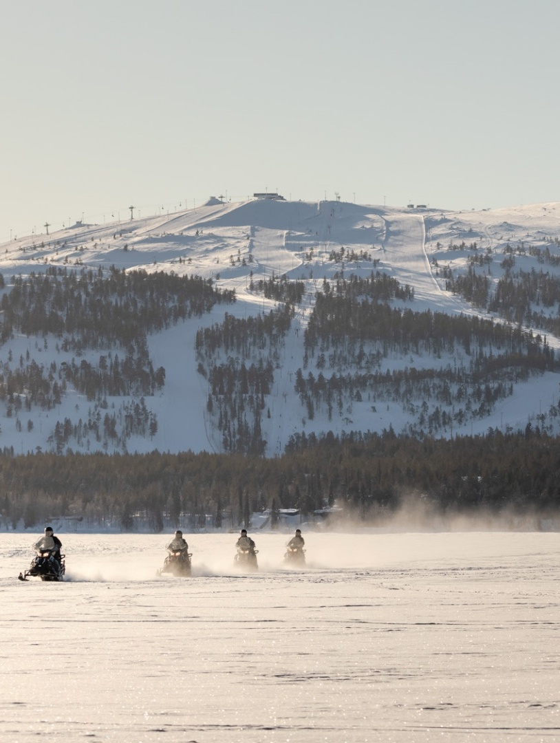 People riding snow scooters, with a large hill coated in snow and covered in trees behind them.