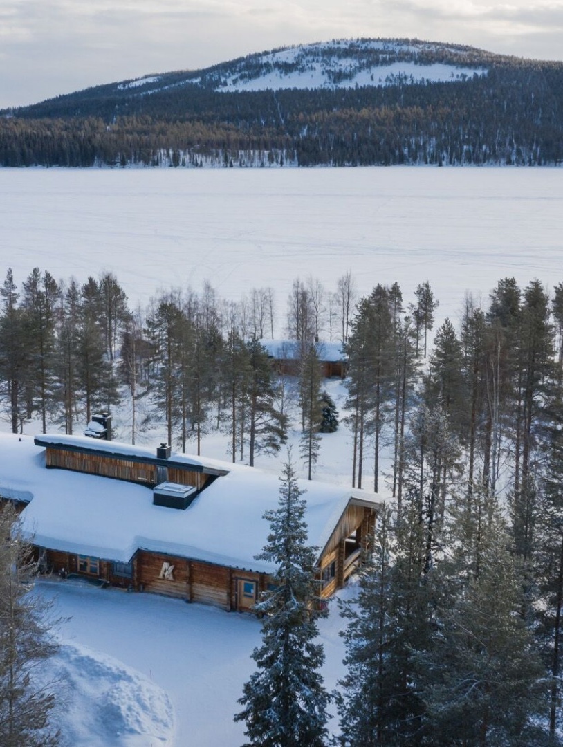 A snow-covered timber lodge surrounded by trees, with other lodges and the lake in the background.