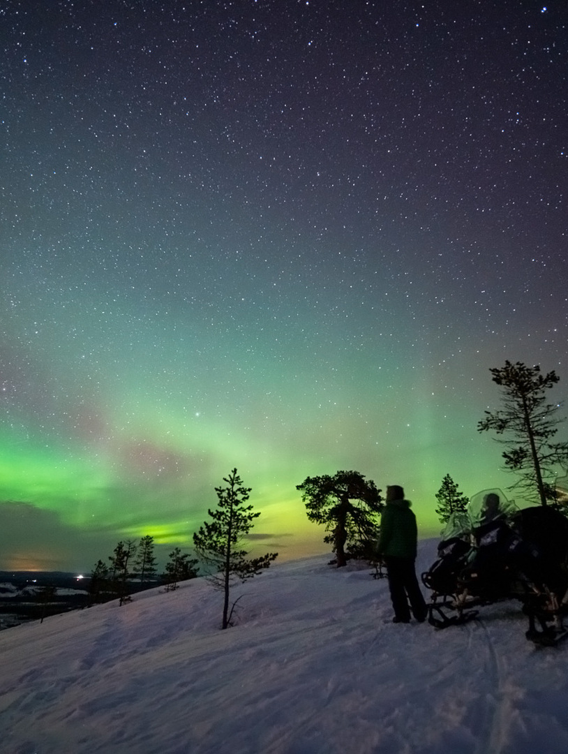 Green and blueish northern lights visible above a snowy hill.