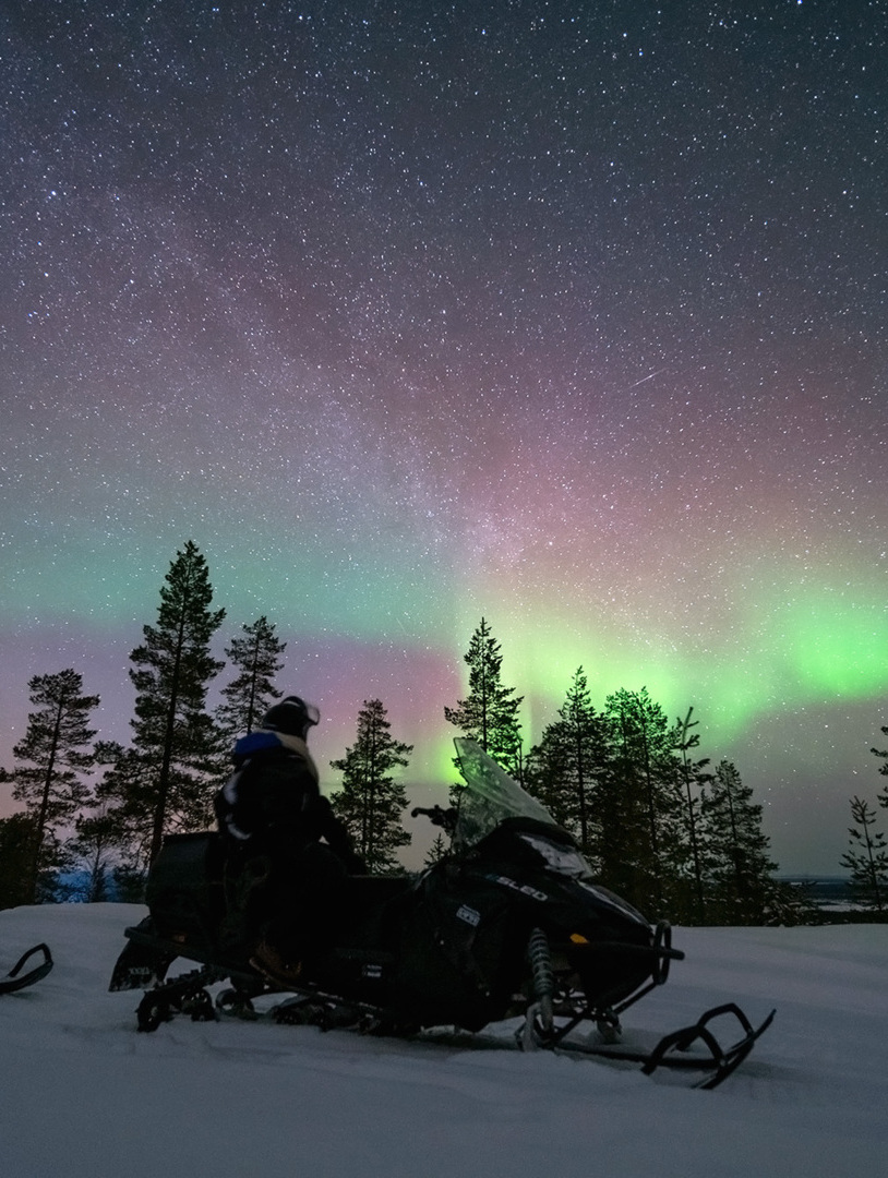 Guests on snowmobiles under a starry sky with northern lights above a snowy forest landscape