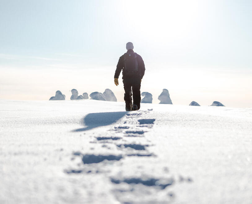 A man hiking through deep snow towards the bright sun light in the distance.