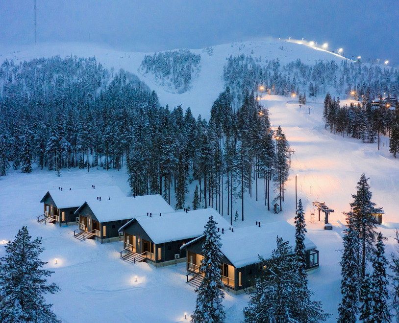 Snow-covered modern cabins with warm interior lights near a ski slope surrounded by forested hills at dusk