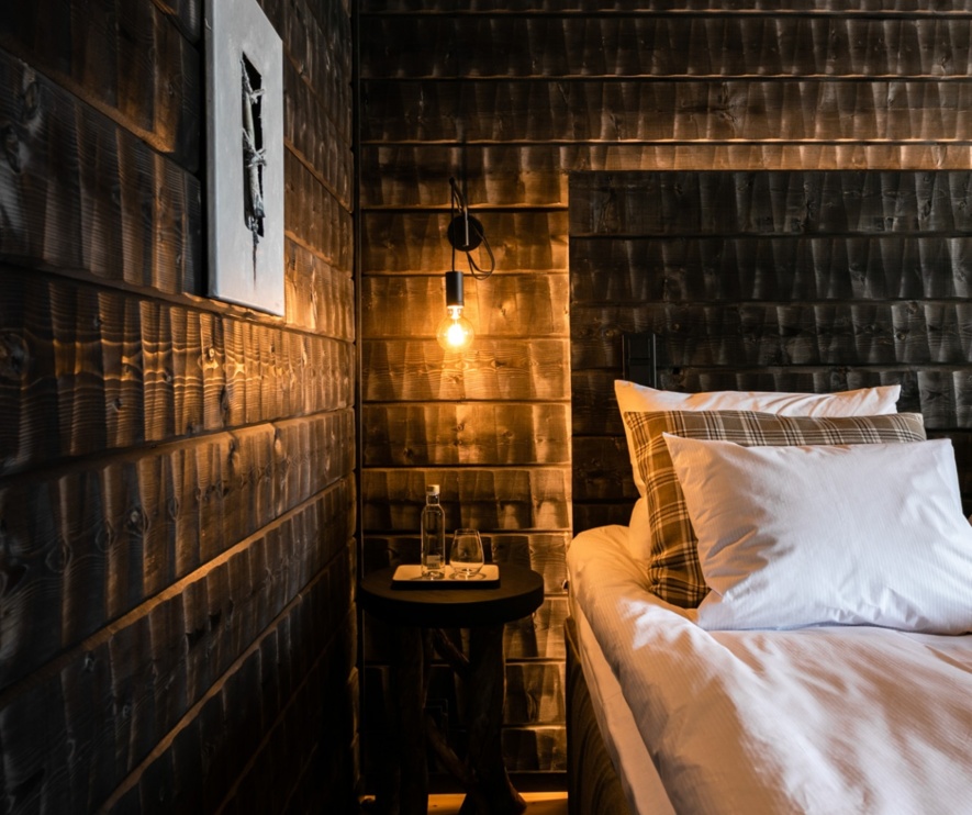 A bedroom with wooden walls, illuminated by golden light from a single lamp above the nightstand.