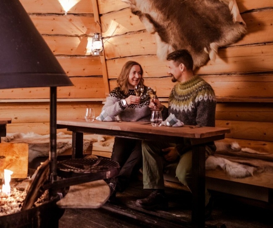A couple sitting at a table in a wooden hut with a fireplace in the middle.