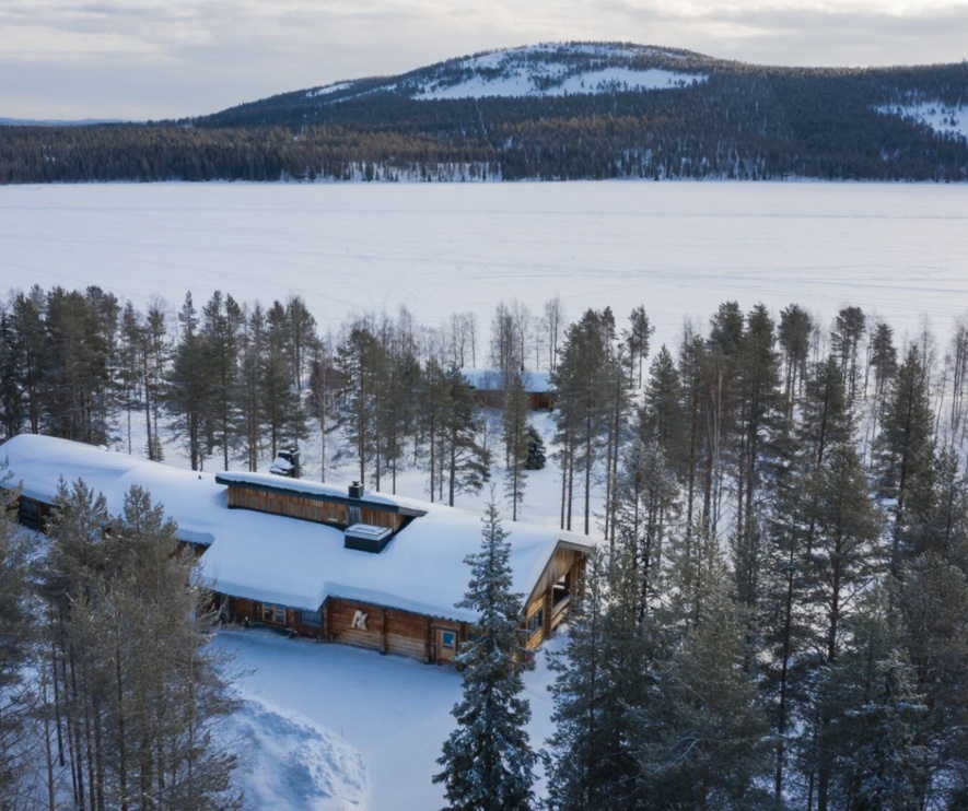 A snow-covered timber lodge surrounded by trees, with other lodges and the lake in the background.