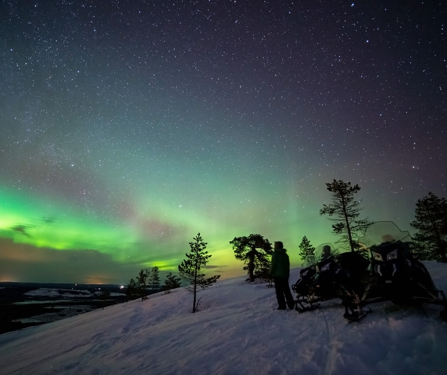 Green and blueish northern lights visible above a snowy hill.