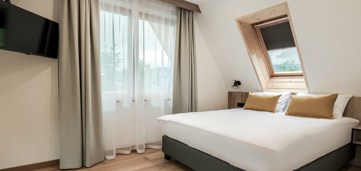 Dwuosobowe łóżko przy oknie, z którego roztacza się widok na malownicze Zakopane i okoliczne góry.