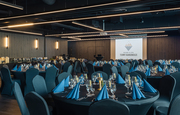 Eleganter Bankettsaal des Hotels, vorbereitet für Gala: runde Tische, dunkelblaue Servietten, glänzendes Glas, Buffet und Projektionsleinwand.