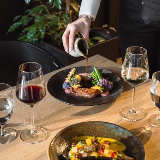 Kelner nalewa sos na podsmażoną kaczkę z sezonowymi warzywami, kieliszki wina przy eleganckim stoliku restauracji