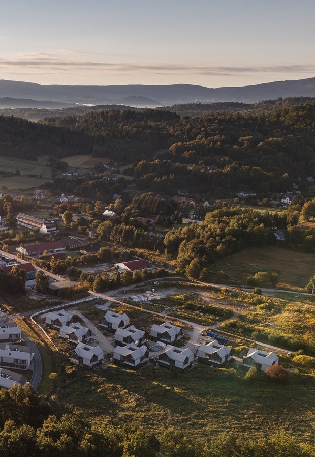 Ośrodek z nowoczesnymi domkami i hotelem w lesie, parking, mgła nad pagórkami i górami o wschodzie.