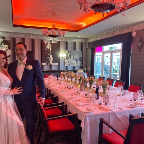 Bride and groom beside a long, elegantly set reception table with flowers, red-upholstered chairs and warm ceiling lighting