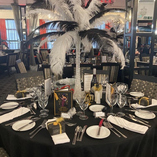 Black-clothed gala table with tall crystal vase topped by black-and-white ostrich feather centerpiece, place settings, menus and wine.