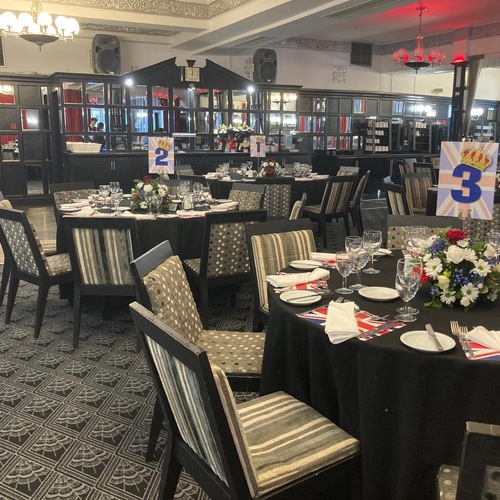 Elegant hotel banquet room set for dinner: round tables with black linens, floral centerpieces, Union Jack table numbers, glassware, chandeliers.
