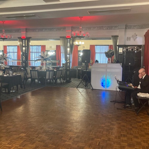 Banquet room with parquet dance floor, set dining tables with feather centerpieces, chandeliers, red curtains and DJ booth.