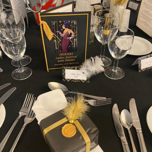 Art Deco gala place setting on black cloth: 'HERMES Ladies Festival Gala Dinner' menu, feathered place card, gold-sealed favor and glassware.