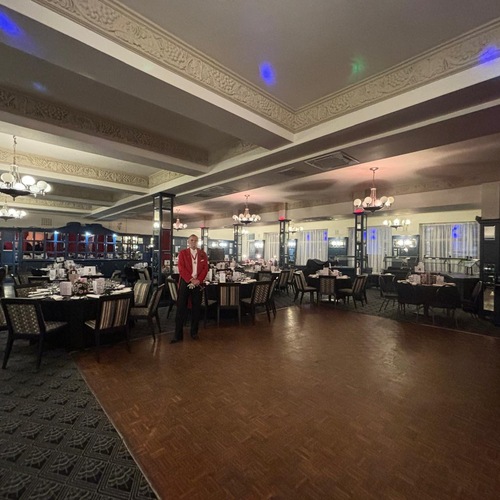 Classic hotel ballroom with ornate coffered ceiling, chandeliers, set round tables and parquet dance floor; a server in a red jacket.