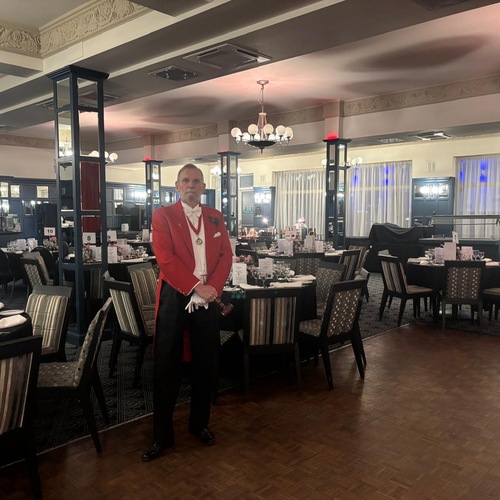Elegant hotel banquet room with set tables and chandeliers; a man in a red tailcoat and white gloves stands holding a wooden gavel.