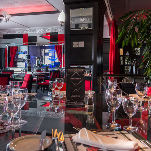 Elegant, intimate hotel restaurant with red-and-black decor, glass-top tables set with wine glasses, silverware and chandelier.