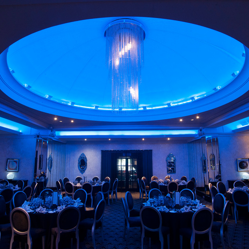 Elegant hotel banquet hall with glowing blue domed ceiling, cascading crystal chandelier and round tables set for a formal dinner.