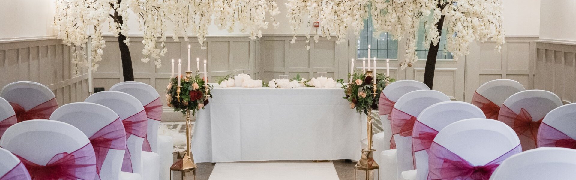 Indoor wedding aisle with white blossom trees framing the head table, white chairs with magenta sashes, candles and lanterns.