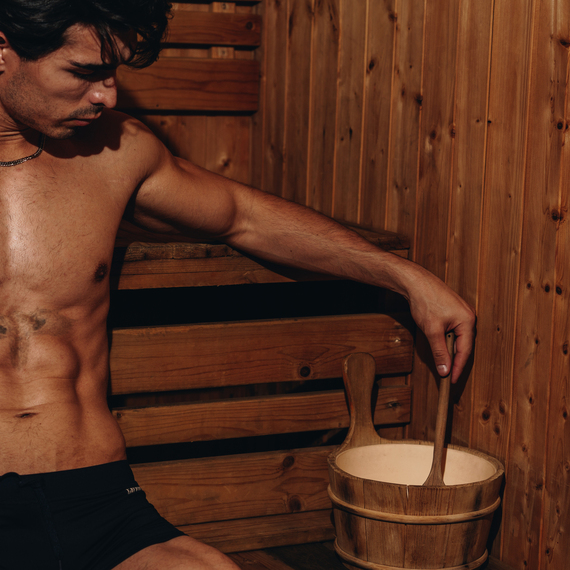 Shirtless guest relaxing in a cedar sauna, seated on wooden benches, stirring water in a wooden bucket with a ladle.