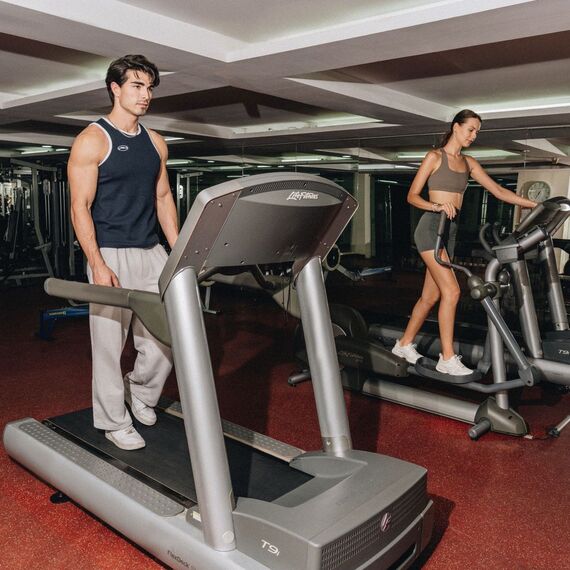 Hotel gym with treadmills and ellipticals, mirrored wall; man on treadmill and woman using elliptical.