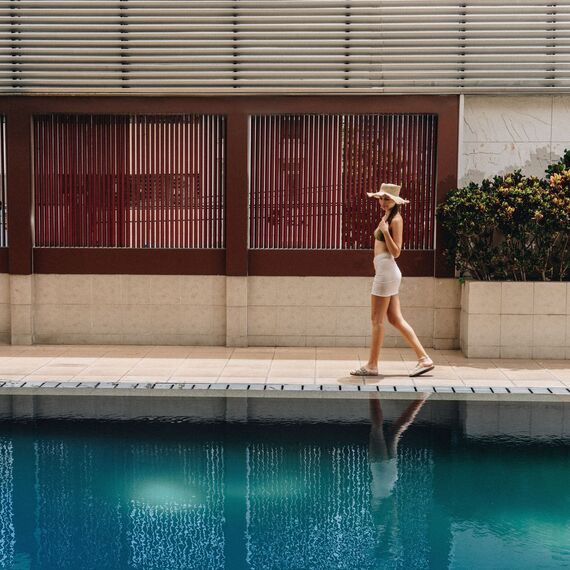 Guest strolling along turquoise hotel pool in sunhat and swimwear, modern slatted privacy wall, lounger and tropical shrubs.