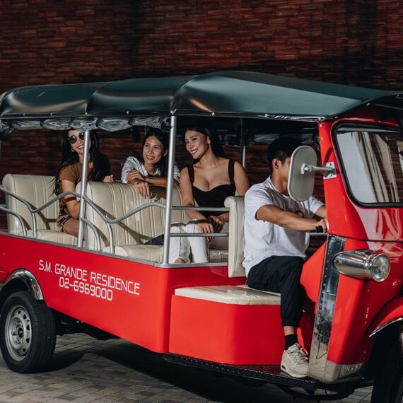 Red hotel tuk-tuk shuttle with driver and three smiling guests seated on cream benches outside a brick-clad residence