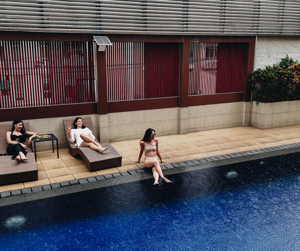 Three women relaxing at hotel pool on tiled deck, two on wicker loungers with drink and fruit, one dipping feet in the water.