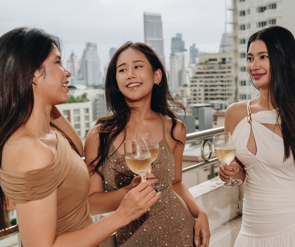 Three women in cocktail dresses toasting with white wine on a hotel rooftop terrace overlooking a city skyline.