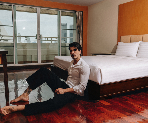 City-view hotel room with large bed, white linens, wood headboard and balcony; guest relaxed, seated on hardwood floor.
