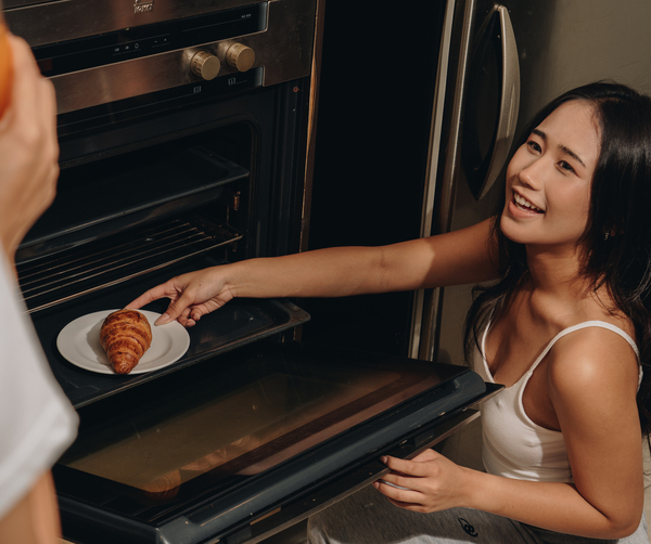Smiling guest removes a golden croissant from a stainless-steel oven in a suite kitchenette as another offers orange juice