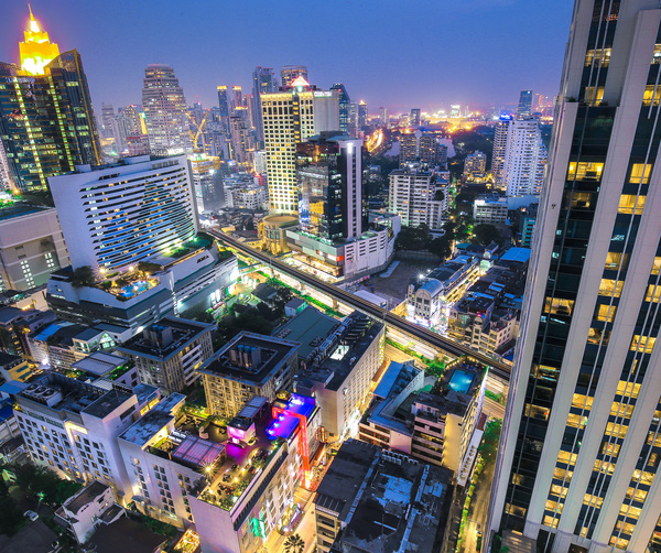 Night city skyline viewed from a hotel, lit high-rises, rooftop pools, glowing guest rooms and an elevated rail.