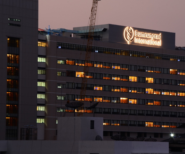 Multi-story international hospital at dusk with rows of warm-lit windows and a glowing rooftop Bumrungrad International sign