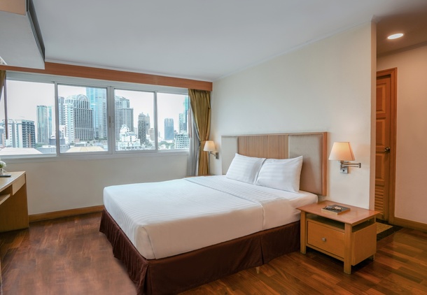 Hotel room with queen bed in white linens, wooden floors, bedside lamps, desk and large window with city skyline view.