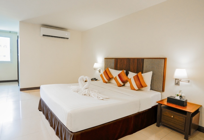 Bright modern hotel room with king bed, wooden headboard, orange striped cushions, towel swans, bedside lamps and wall AC.