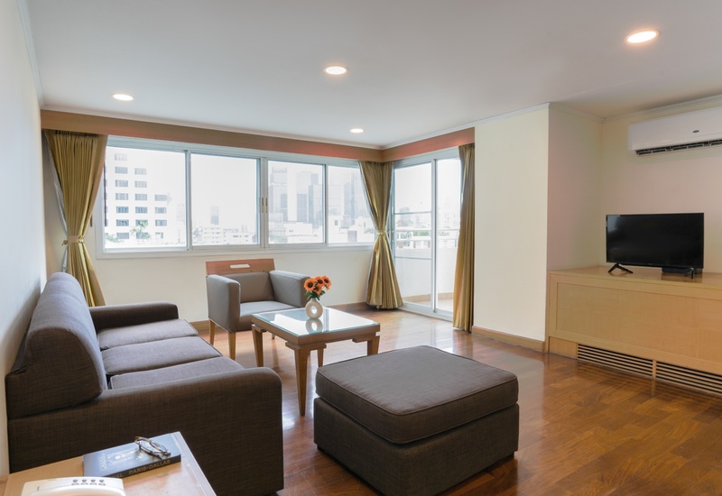 Hotel suite living area with gray sofa, ottoman, armchair, coffee table with flowers, TV, AC and city-view windows.