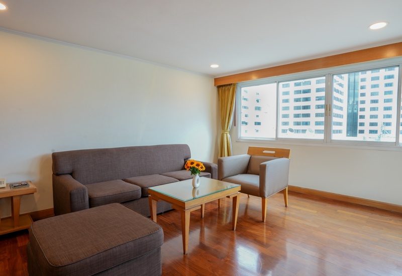 Hotel suite seating with brown sofa, armchair, ottoman, glass coffee table with sunflowers and city-view windows