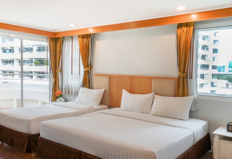 Bright twin room with two white-linen beds, wooden headboard, golden drapes, bedside table, and city views.