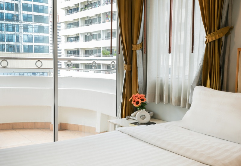 Bright hotel room with king bed, sheer curtains and gold drapes, balcony with city view, bedside table with flowers.