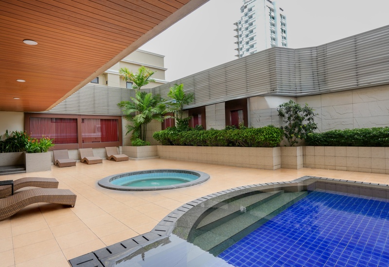 Hotel outdoor pool area with blue-tiled pool and round hot tub, curved wicker loungers, potted palms and city view.