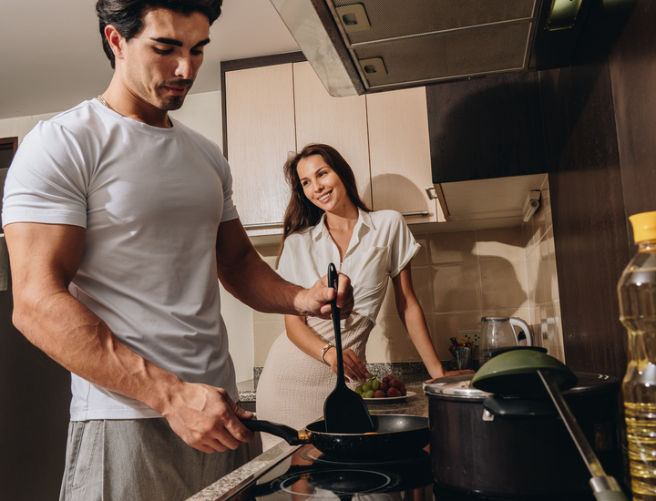 Couple preparing breakfast in a modern suite kitchenette, man frying eggs on stovetop, woman smiling beside fruit on granite counter