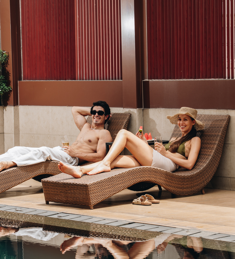 Couple relaxing on wicker pool loungers by a private hotel pool, sipping drinks and enjoying sun on a tiled terrace.