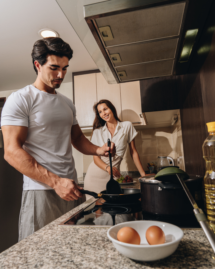 Couple preparing breakfast in a modern suite kitchenette, man frying eggs on stovetop, woman smiling beside fruit on granite counter