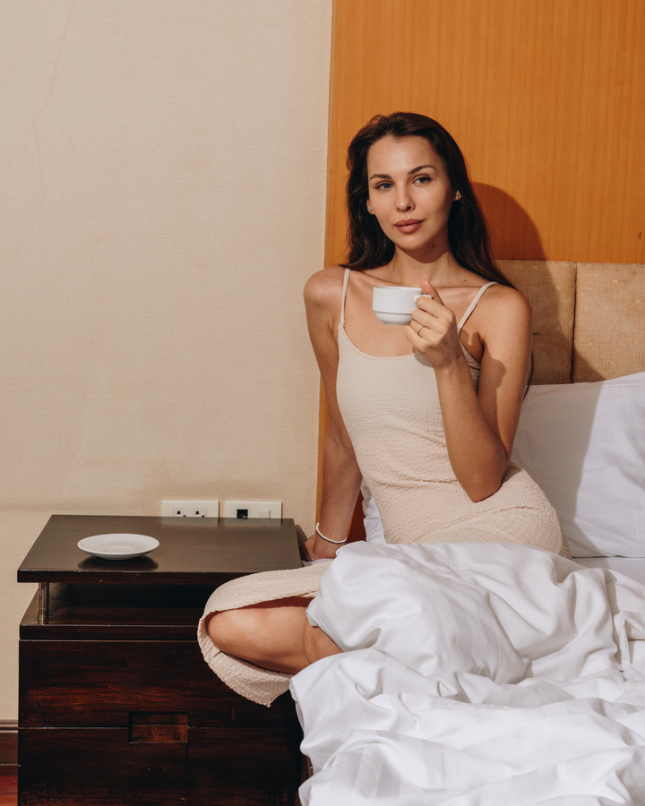 Woman in a light slip dress sitting on a hotel bed, sipping coffee beside a wooden nightstand with white bedding.