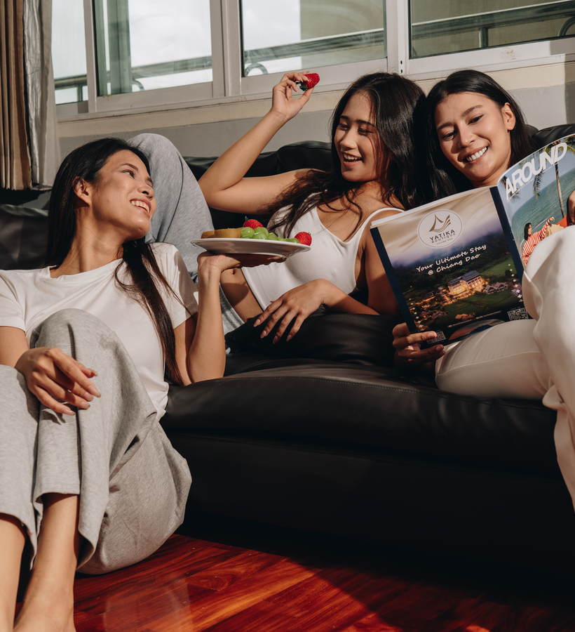 Three friends lounging on a hotel sofa by a sunlit window, sharing fresh fruit and browsing the hotel's brochure