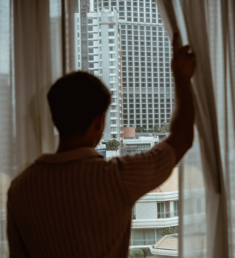 Guest silhouette pulls sheer curtains in a hotel room, revealing a city skyline and neighboring high-rise hotel through a large window